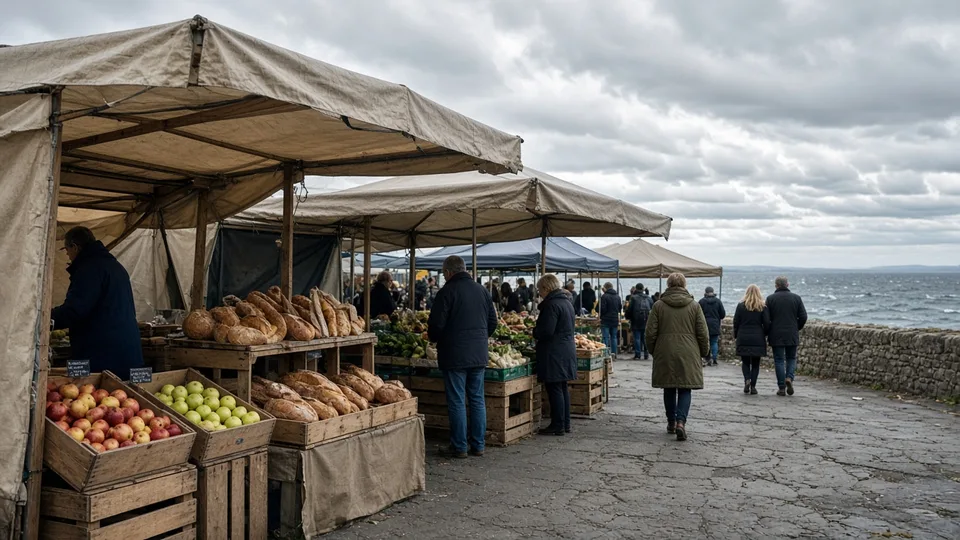 Outdoor food market stalls on a windy seafront, with bread and fruit under canvas and shoppers in coats.